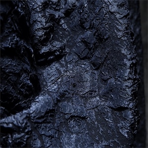 May include: Detailed shot of a dark navy blue fabric with a textured surface. The fabric has a crinkled, uneven appearance, creating a play of light and shadow. The textile's rich colour and texture suggest its use in clothing or interior design.