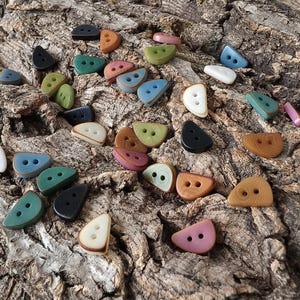 May include: A collection of semi-circular buttons in various colors including black, white, green, blue, brown, and pink. Each button features two small holes in the center and a smooth, rounded edge, scattered across a textured, rough surface.