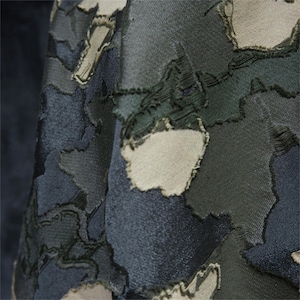 Op de afbeelding: Close-up van een stof met een camouflagepatroon. De stof heeft vlekken in verschillende tinten groen, grijs en beige, waardoor een getextureerd, gelaagd effect ontstaat. De randen van de vlekken zijn gerafeld, wat bijdraagt aan het unieke ontwerp van de stof.