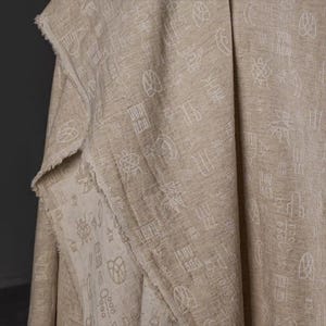 May include: A beige fabric with a visible woven texture featuring a repeating pattern of white line-drawn symbols and geometric shapes. The material has a raw, frayed edge along the folded section, showcasing the natural weave of the textile.