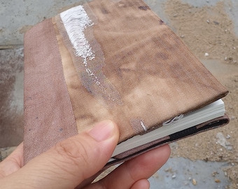 May include: A small, handmade journal with a brown fabric cover. The cover has a textured appearance with a lighter brown spine and a white, textured design. The journal is held in a hand.