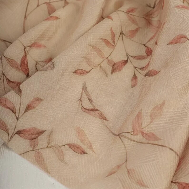 Op de afbeelding: Close-up van een beige stof met een delicaat patroon van bladeren en takken in roze en bruine tinten. De stof heeft een subtiele rasterachtige textuur, wat een zachte, natuurlijke uitstraling cre&euml;ert. Ideaal voor knutselen of kleding.