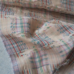Puede incluir: Primer plano de una tela beige con diseño de patchwork. La tela presenta un patrón de cuadros en rojo, azul y rosa, con bordes deshilachados. La tela está doblada, mostrando la textura y los detalles.