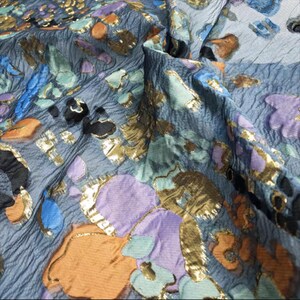 May include: Close-up of a textured fabric with a floral pattern. The fabric is a muted blue-grey with gold, orange, purple, and teal floral accents. The fabric has a crinkled appearance, adding depth and texture to the design.