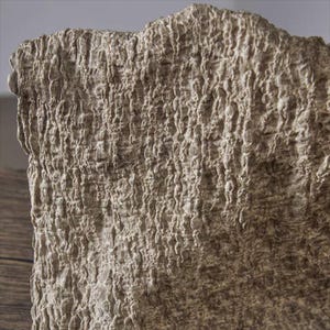 May include: Close-up of a textured, beige fabric with a rough, uneven surface. The fabric has a crinkled appearance, with vertical lines and a slightly wavy top edge. The neutral color and texture suggest a natural fiber.