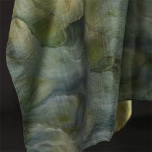 May include: A draped fabric with a watercolor-like pattern in shades of green, blue, and cream. The fabric has a textured appearance, with visible woven threads. The overall effect is soft and flowing, suggesting a scarf or shawl.