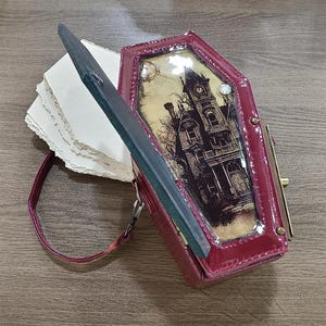 Puede incluir: Bolso de mano en forma de ataúd burdeos con una ilustración de una casa embrujada. El bolso tiene una correa larga, un cierre de broche y una tapa con bisagras que revela una pila de papel. El bolso es de material brillante.