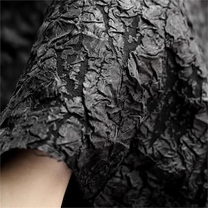 以下が含まれることがあります： しわのある、層状の外観の黒いテクスチャード加工された生地のクローズアップ。生地は、さまざまな黒とグレーの色合いを持つ、ざらざらした、ほぼ樹皮のような質感です。素材はドレープまたは折りたたまれているように見え、影と奥行きを作り出しています。