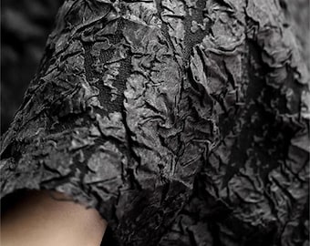 May include: Close-up of a textured black fabric with a crinkled, layered appearance. The fabric has a rough, almost bark-like texture, with varying shades of black and gray. The material appears to be draped or folded, creating shadows and depth.
