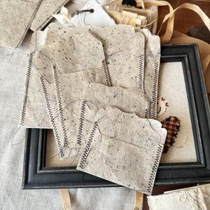 Könnte beinhalten: Eine Sammlung handgefertigter Papierumschläge mit einer strukturierten, gesprenkelten Optik. Jeder Umschlag ist mit schwarzem Faden an den Rändern genäht. Die Umschläge sind in der Nähe eines gerahmten Kunstwerks mit schwarzem Rahmen angeordnet.
