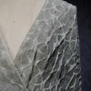 May include: Close-up of a gray fabric with a textured, patterned design. The fabric appears to be draped, with a V-neckline visible. The texture has a raised, irregular pattern, creating a unique visual effect.