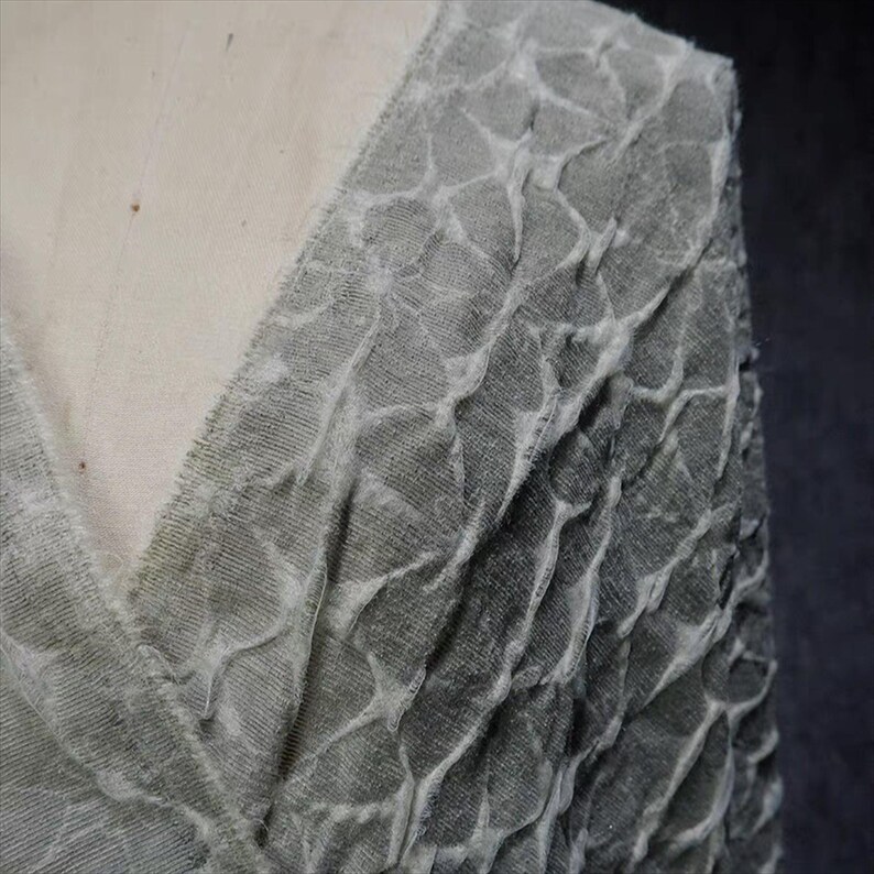 May include: Close-up of a gray fabric with a textured, patterned design. The fabric appears to be draped, with a V-neckline visible. The texture has a raised, irregular pattern, creating a unique visual effect.
