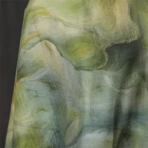 May include: A close-up of a fabric with a watercolor effect. The fabric features a blend of green, yellow, and blue hues, creating a soft, flowing pattern. The texture appears slightly sheer, with visible woven details. The overall effect is reminiscent of a landscape.