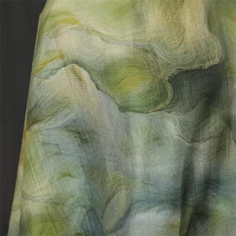 May include: A close-up of a fabric with a watercolor effect. The fabric features a blend of green, yellow, and blue hues, creating a soft, flowing pattern. The texture appears slightly sheer, with visible woven details. The overall effect is reminiscent of a landscape.