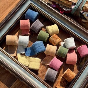 May include: A collection of small, rolled fabric ribbons in various colors including blue, pink, yellow, green, grey, and tan, arranged inside a dark wooden picture frame. The ribbons feature frayed edges and a textured, matte appearance.