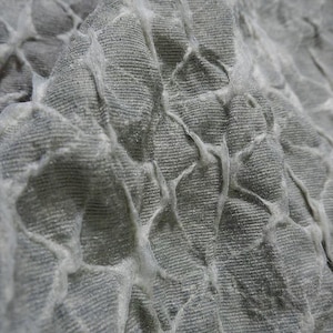 May include: Close-up of a textured, gray fabric with a raised, web-like pattern. The fabric appears to be soft and pliable, with a subtle sheen. The raised pattern creates a three-dimensional effect, adding visual interest to the textile.
