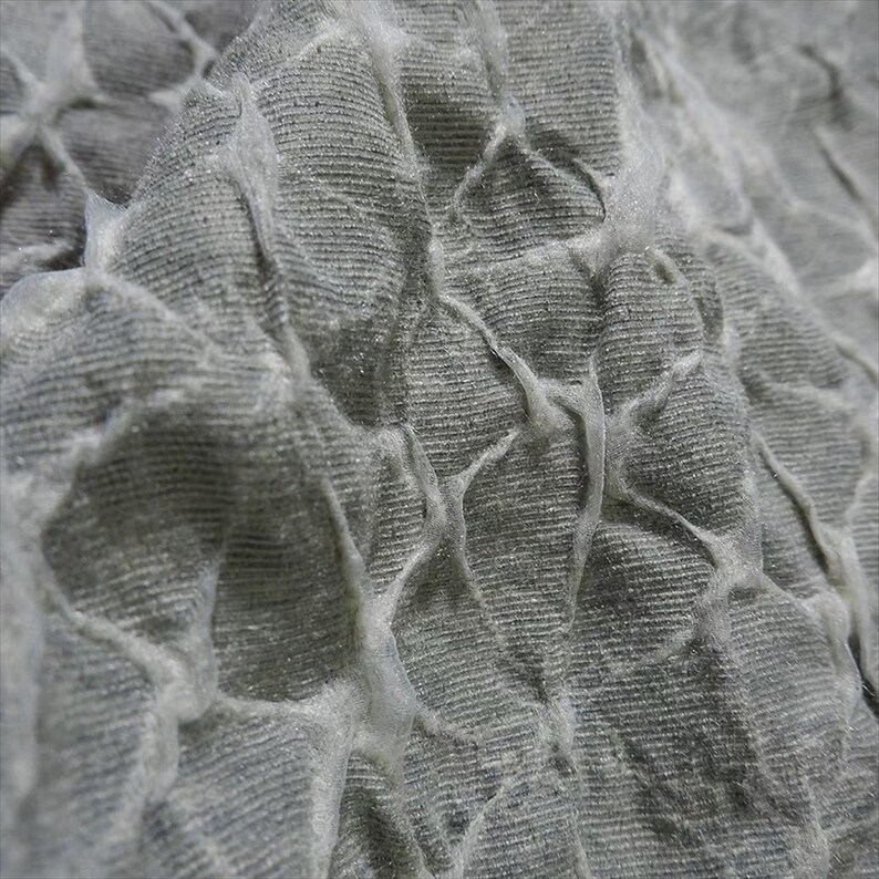 May include: Close-up of a textured, gray fabric with a raised, web-like pattern. The fabric appears to be soft and pliable, with a subtle sheen. The raised pattern creates a three-dimensional effect, adding visual interest to the textile.