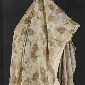May include: A lightweight fabric drape featuring an all-over botanical print with various leaf shapes in shades of tan, beige, and muted brown. The textile displays a natural, organic pattern with overlapping foliage silhouettes across the surface.
