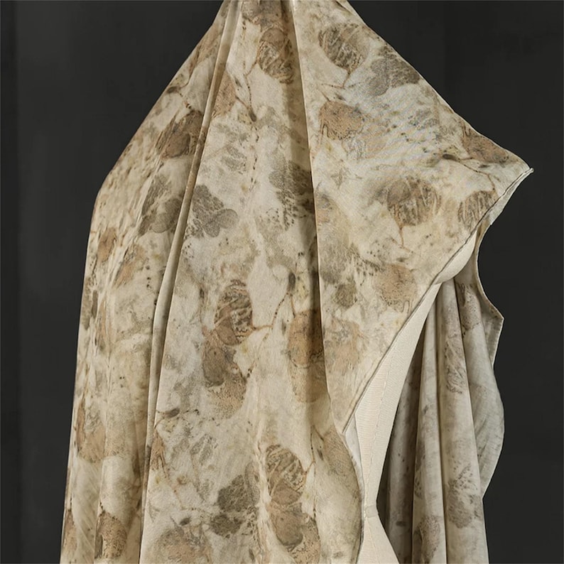 May include: A lightweight fabric drape featuring an all-over botanical print with various leaf shapes in shades of tan, beige, and muted brown. The textile displays a natural, organic pattern with overlapping foliage silhouettes across the surface.