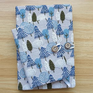May include: Two embroidered fabric book covers, one large and one small, with a light gray background and a pattern of blue, white, and green trees. The larger cover has a button closure. The smaller cover is on top of the larger one.