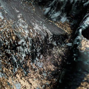 May include: Close-up of a textured fabric with a dark teal base, overlaid with shimmering gold and silver accents. The fabric has a luxurious, flowing appearance, with visible folds and subtle variations in color and texture.