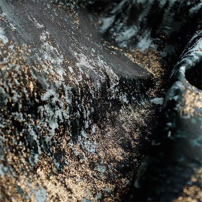 May include: Close-up of a textured fabric with a dark teal base, overlaid with shimmering gold and silver accents. The fabric has a luxurious, flowing appearance, with visible folds and subtle variations in color and texture.