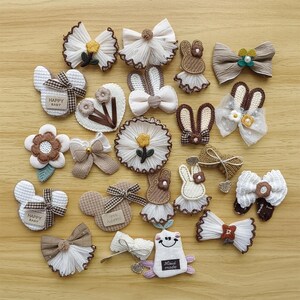 May include: A collection of assorted hair accessories, including bows, flower shapes, and bunny designs. The accessories feature various textures, colors, and embellishments, with some pieces displaying the text "HAPPY BABY" and "Hand made".