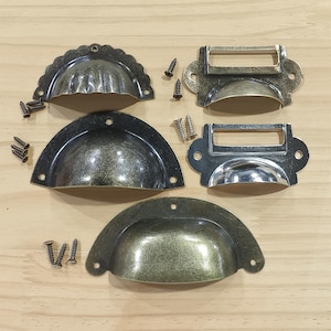 Puede incluir: Un surtido de tiradores y portatarjetas de cajón de estilo antiguo en varios acabados metálicos. Los tiradores tienen forma de media luna y los portatarjetas tienen una ranura rectangular para etiquetas. Se incluyen varios tornillos.