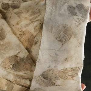 May include: A lightweight fabric scarf featuring an off-white base color with a botanical print pattern in shades of brown, tan, and muted gray. The textile displays organic leaf shapes and natural textures throughout the surface of the material.