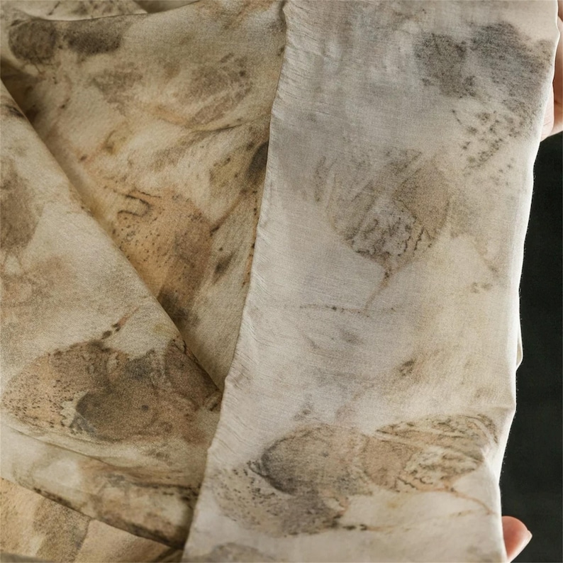 May include: A lightweight fabric scarf featuring an off-white base color with a botanical print pattern in shades of brown, tan, and muted gray. The textile displays organic leaf shapes and natural textures throughout the surface of the material.