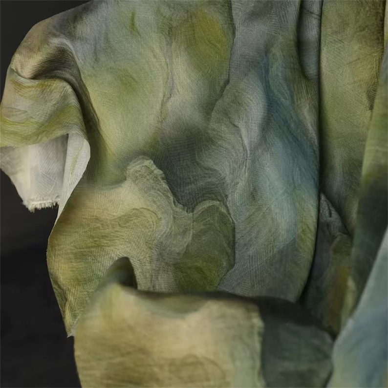 May include: Close-up of flowing fabric in shades of green, blue, and cream. The material has a subtle, rippled texture, with areas of light and shadow creating depth. The fabric appears lightweight and semi-transparent.