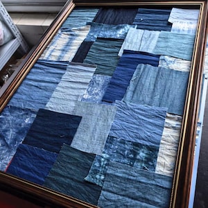 Op de afbeelding: Een rechthoekig wandkunstwerk bestaande uit een collage van diverse stoffen in blauwtinten, variërend van licht hemelsblauw tot diep marineblauw. De stoffen hebben verschillende texturen en patronen en zijn gevat in een donkerbruine lijst.