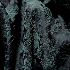 May include: Close-up of a dark green and black floral brocade fabric. The fabric has a raised, textured pattern of flowers and leaves. The folds and shadows create depth and dimension, highlighting the intricate design. Ideal for apparel or home decor.