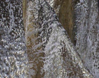 May include: Close-up of a shimmering fabric with a textured surface. The fabric features a blend of silver, gold, and white tones, creating a luxurious and elegant appearance. The material appears to be draped, showcasing its flowing quality.