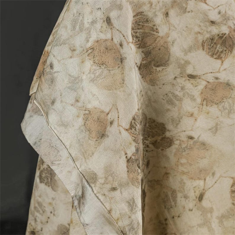 May include: A light-colored fabric garment featuring an all-over botanical print with various leaf shapes in shades of tan, beige, and muted brown. The textile displays a soft, draped texture with visible seams along the edge and a folded corner.