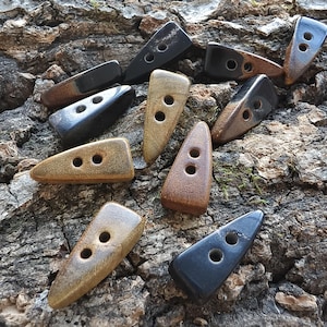 May include: A collection of ten triangular toggle buttons in various shades of brown, tan, and black. Each button features a smooth, polished finish and two circular holes centered along the length for attachment to garments or accessories.