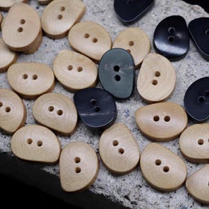 May include: A collection of wooden buttons in various shades of light brown, dark green, and black. The buttons are oval-shaped with two holes for sewing. The buttons are arranged on a textured, gray surface.
