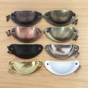 Puede incluir: Un surtido de siete tiradores de metal en forma de taza en varios acabados, incluyendo bronce, cobre, negro, plata, oro y blanco. Cada tirador viene con tornillos pequeños para la instalación. Los tiradores están dispuestos sobre una superficie de madera clara.