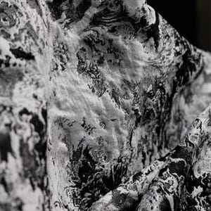 Puede incluir: Primer plano de una tela con un patrón abstracto y arremolinado en tonos grises y negros. La tela tiene una apariencia texturizada, con áreas de luz y sombra que crean profundidad e interés visual. El diseño se asemeja a un efecto marmolado o acuarela.