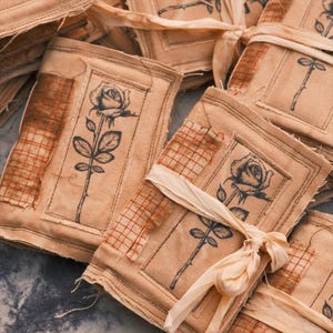 May include: A collection of handmade, beige fabric pouches, each featuring a black rose illustration. The pouches are adorned with patches of red grid-patterned fabric and tied with cream-colored ribbons. The overall aesthetic is rustic and artistic.
