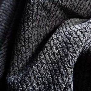 Könnte beinhalten: Nahaufnahme eines dunkelgrauen Stoffes mit einem strukturierten, gewebten Muster. Der Stoff hat einen subtilen Glanz, und das Gewebe erzeugt eine visuelle Textur. Die Falten und Schatten verleihen dem Bild Tiefe und heben das komplizierte Design hervor.