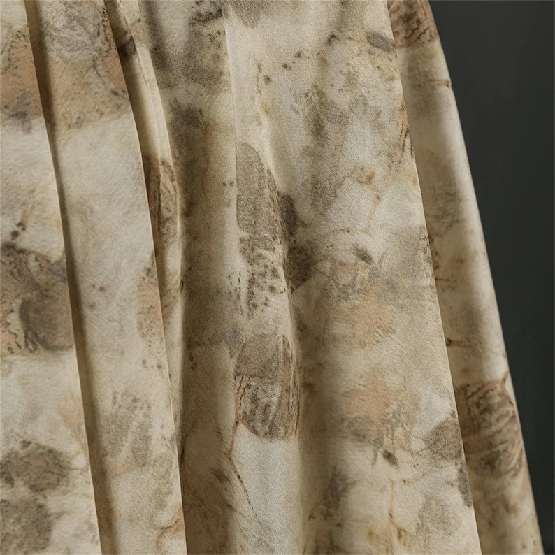 May include: A lightweight fabric featuring a botanical print with overlapping leaf and foliage patterns in muted shades of beige, tan, and soft brown. The material displays vertical folds and drapes, highlighting the intricate, organic textures.