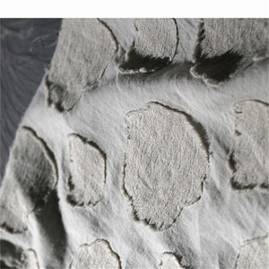 May include: Close-up of a textured, light gray fabric with raised, irregular shapes. The shapes have a rough, almost torn appearance, creating a tactile and visually interesting surface. The fabric's texture and design suggest a focus on textiles and home decor.