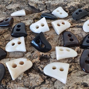 Op de afbeelding: Een verzameling asymmetrische knopen met twee gaten in glanzende zwarte, witte en doorschijnende bruine tinten. Elke knoop heeft een gebogen, driehoekige vorm met gladde, afgeronde randen en een gepolijste afwerking, verspreid over een getextureerd oppervlak.