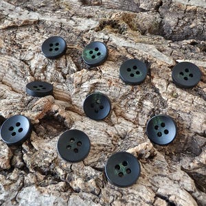 May include: A collection of ten round, matte black buttons, each featuring four holes in the center. The buttons have a subtle, dark green, moss-like texture or patina visible within the recessed areas around the central holes.