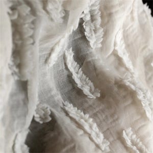 May include: Close-up of a white fabric with a textured, layered design. The fabric features raised, leaf-like patterns that create a sense of depth and dimension. The material appears soft and delicate, with a subtle sheen.