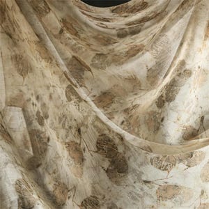 May include: A lightweight, semi-sheer fabric scarf featuring an all-over botanical print of scattered brown and tan leaves on a cream-colored background. The material is draped in soft folds, highlighting the intricate, organic leaf patterns.