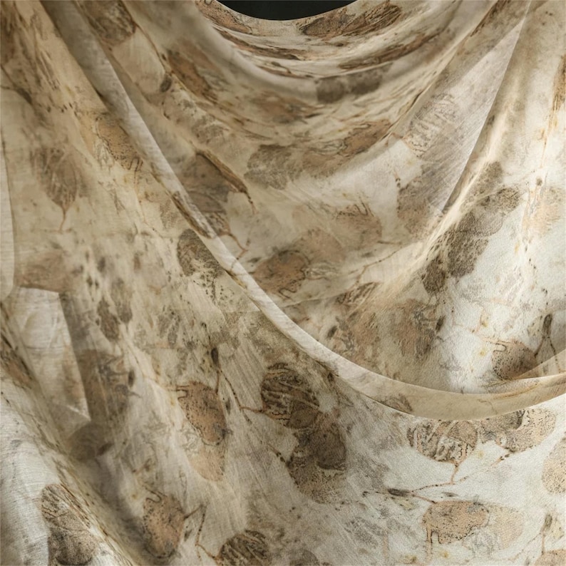 May include: A lightweight, semi-sheer fabric scarf featuring an all-over botanical print of scattered brown and tan leaves on a cream-colored background. The material is draped in soft folds, highlighting the intricate, organic leaf patterns.