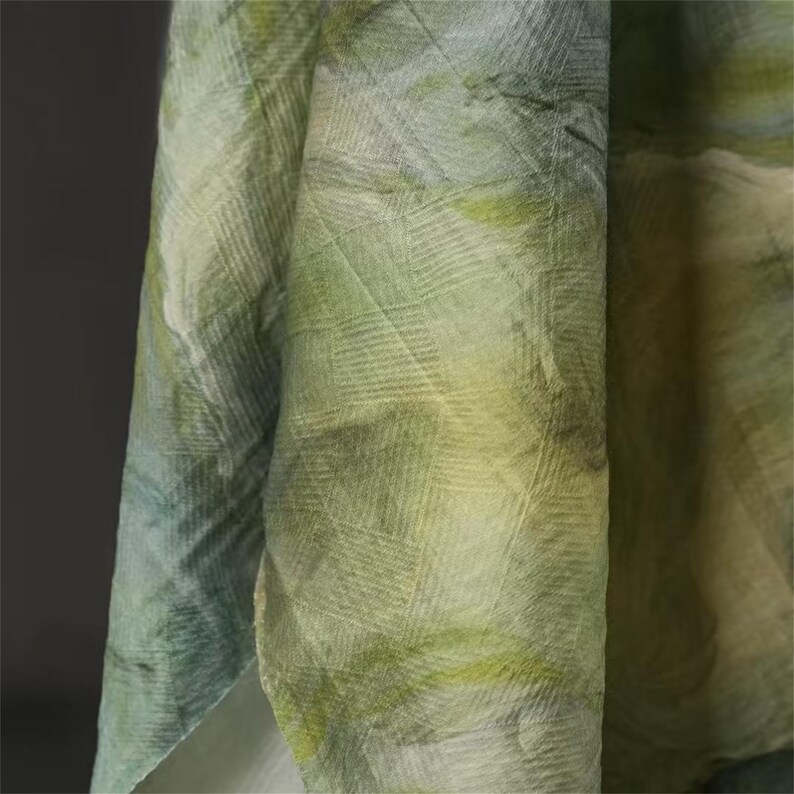 May include: Close-up of a flowing fabric with a watercolor effect. The material features a blend of green, yellow, and blue hues, creating a soft, textured appearance. The fabric is draped, showcasing its lightweight quality and subtle pattern.