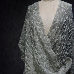 May include: A light gray, textured wrap-style garment with a V-neck design. The fabric has a repeating, geometric pattern, creating a layered effect. The garment is draped over a mannequin, showcasing its shape and texture.